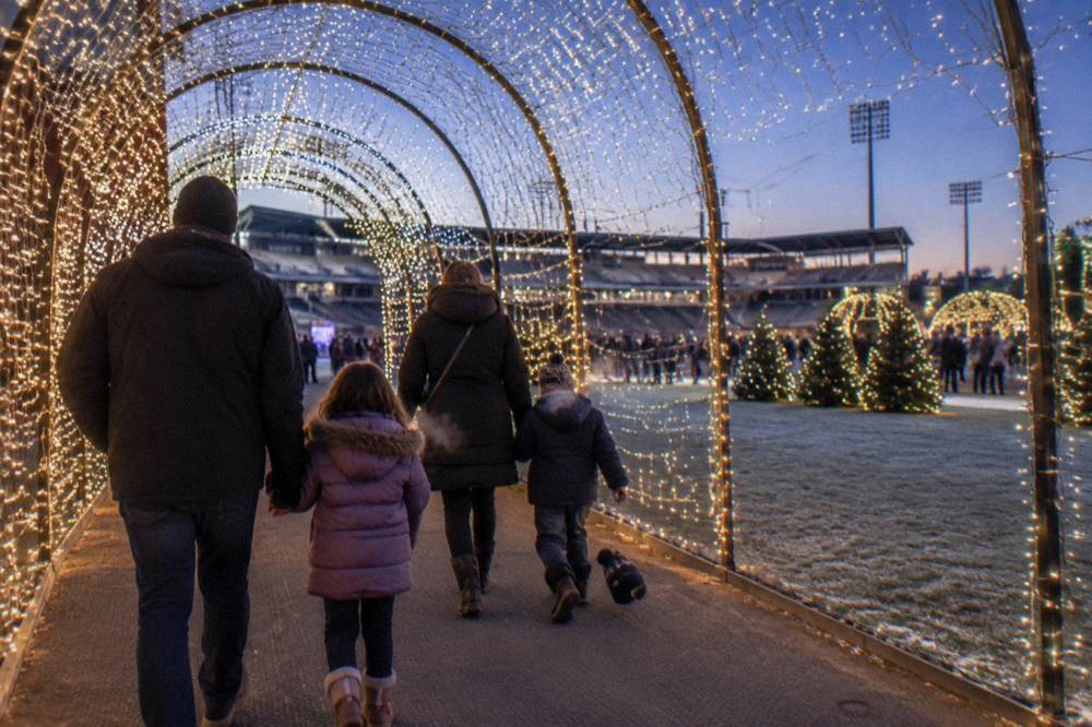 A family walking through a glowing tunnel of string lights at an outdoor holiday event on a cold clear evening.