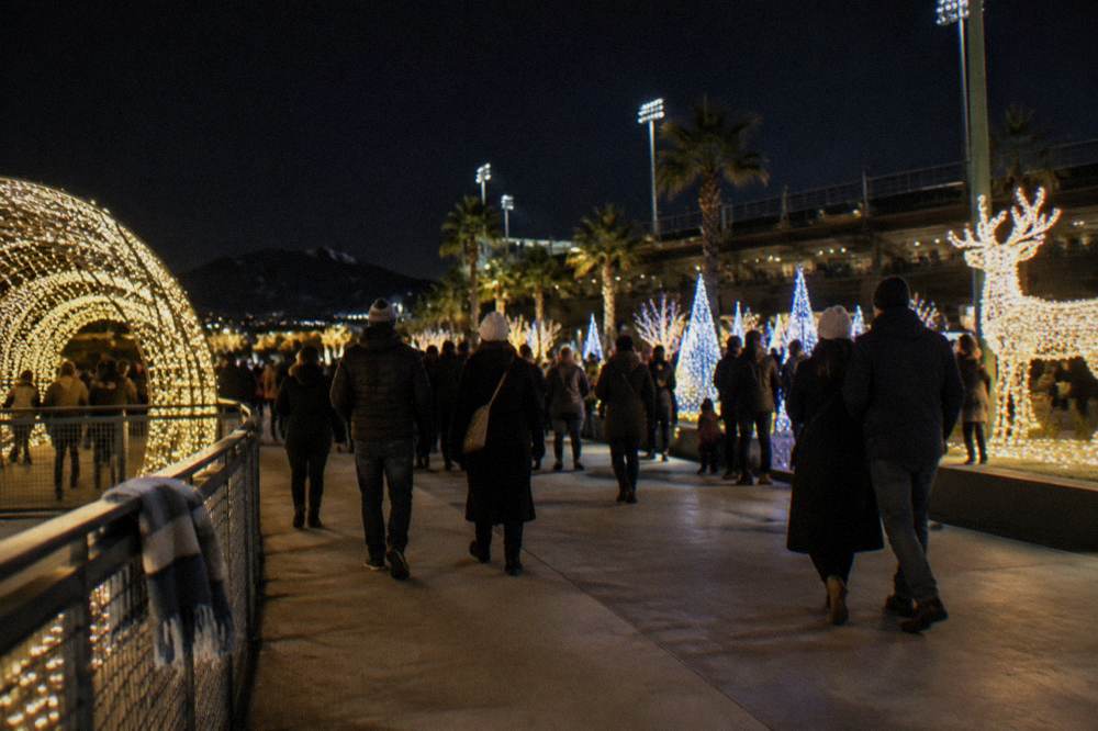 Warmly dressed visitors in coats and hats walking between glowing holiday light displays at an outdoor winter event.