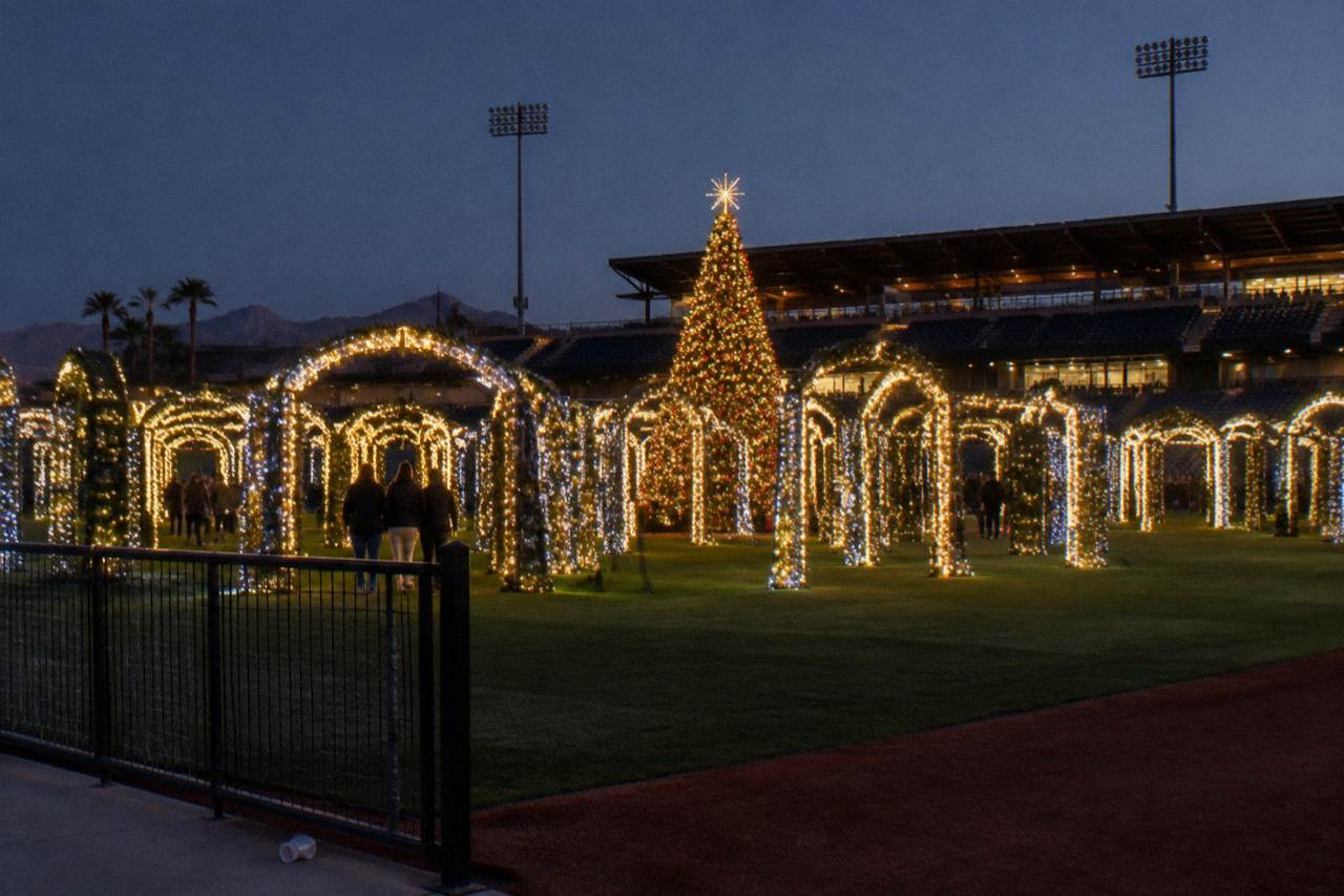 A glowing holiday light maze with illuminated archways and a decorated Christmas tree at an outdoor ballpark venue after dark.