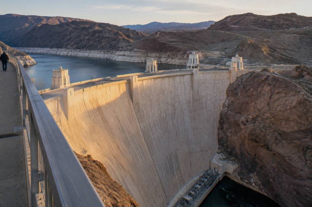 Curved concrete face of a large dam with a steel bridge spanning the canyon above it