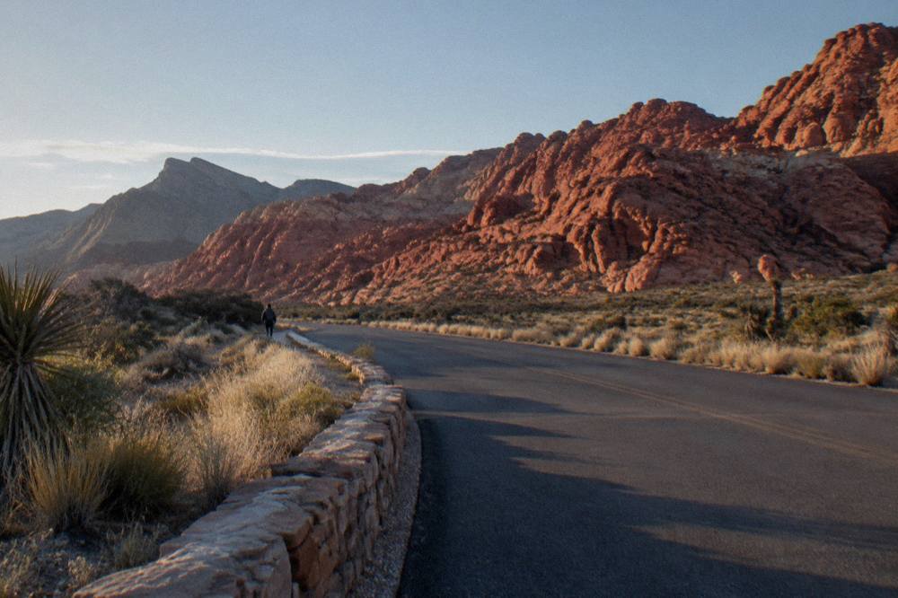 A desert road curving toward layered red sandstone ridges in late afternoon light